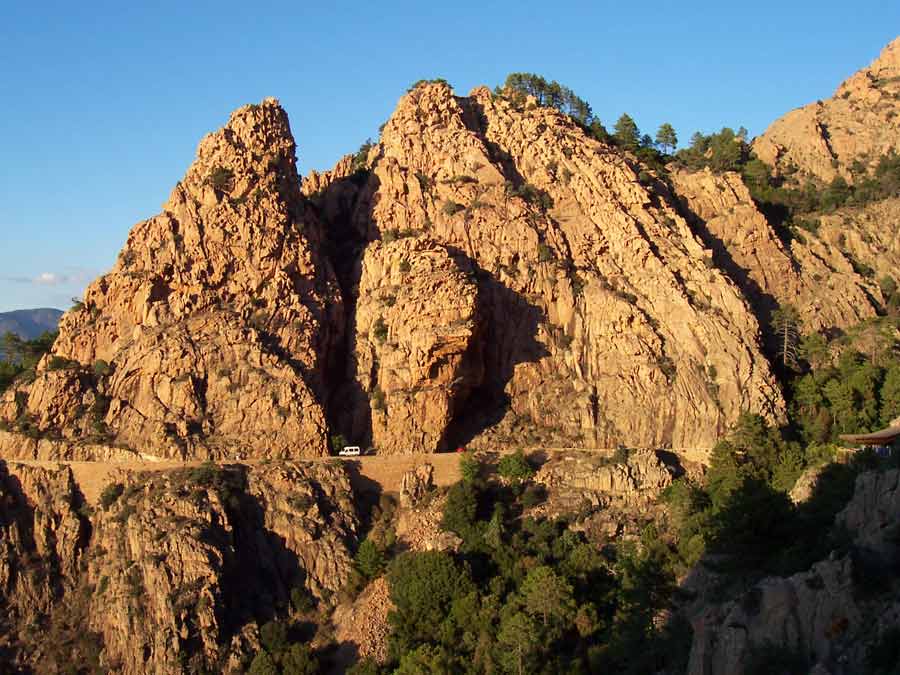 Route étroite serpentant au pied des impressionnantes falaises rouges des calanques de Piana en Corse, avec un véhicule passant entre les formations rocheuses