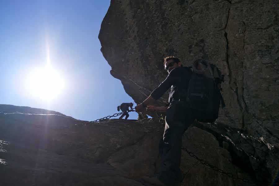 Randonneur agrippé aux chaînes sur la variante alpine du GR20, sous le soleil des aiguilles de Bavella en Corse