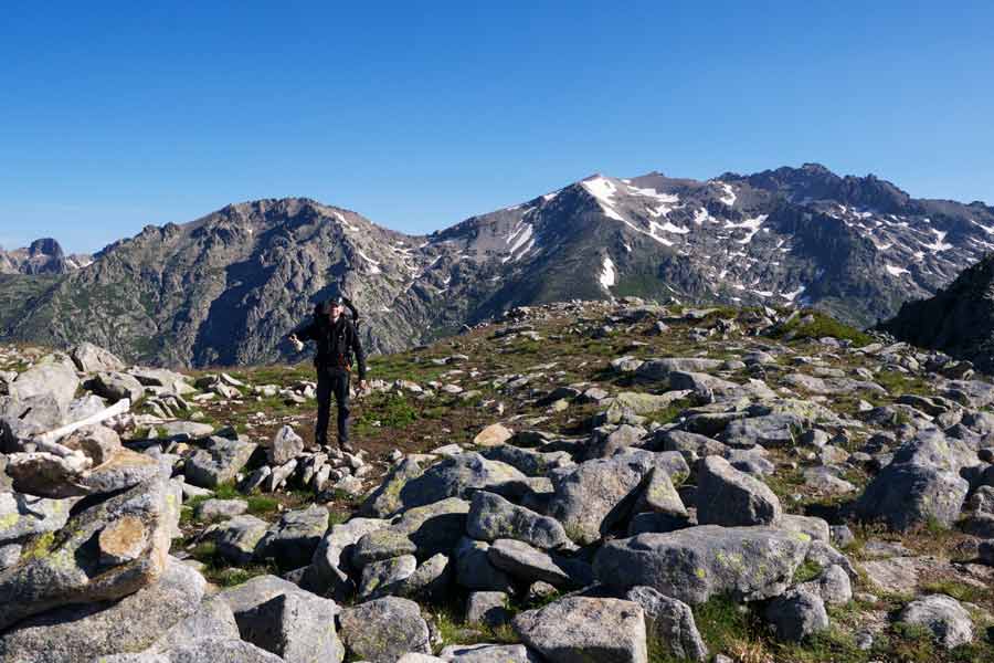 Randonneur au milieu d’un paysage minéral sur le GR20 entre L’Onda et Petra Piana, face aux montagnes enneigées de Corse