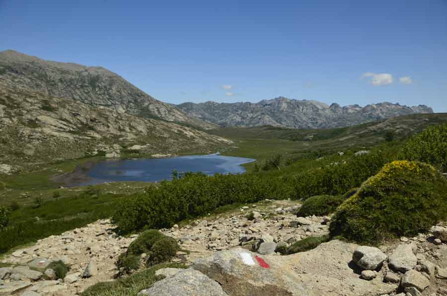 Lac de montagne en Corse entouré de rochers et de végétation avec sommets en arrière-plan